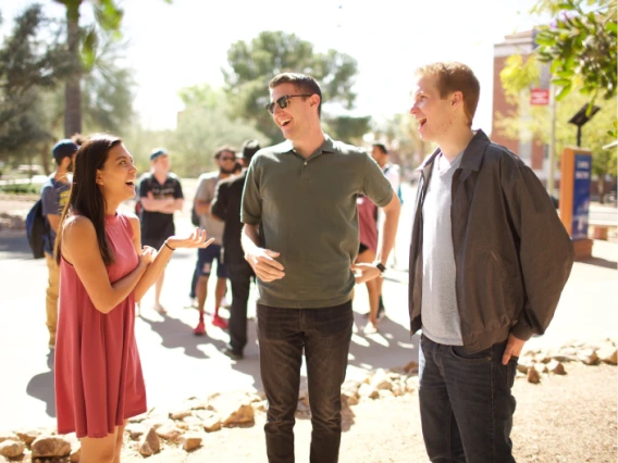 Three students talking outside on campus with more students and school building in the background